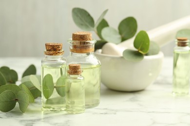 Photo of Bottles of essential oil, eucalyptus leaves, mortar and pestle on white marble table, closeup