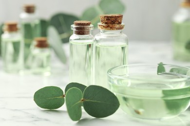 Photo of Bottles of essential oil, bowl and eucalyptus leaves on white marble table, closeup