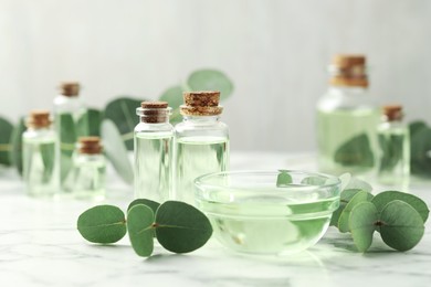 Photo of Bottles of essential oil, bowl and eucalyptus leaves on white marble table, closeup