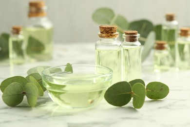Photo of Bottles of essential oil, bowl and eucalyptus leaves on white marble table, closeup