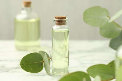 Photo of Bottle of essential oil and eucalyptus leaves on white marble table, closeup