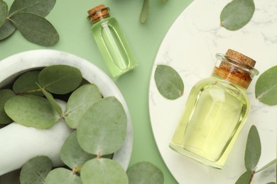 Photo of Bottles of essential oil, eucalyptus leaves, mortar and pestle on green background, flat lay