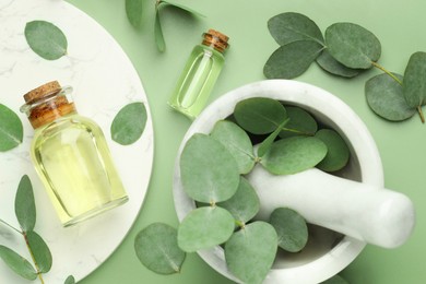 Photo of Bottles of essential oil, eucalyptus leaves, mortar and pestle on green background, flat lay
