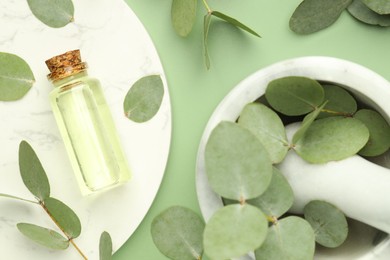 Photo of Bottle of essential oil, eucalyptus leaves, mortar and pestle on green background, flat lay