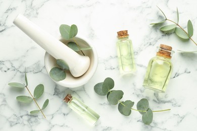 Photo of Bottles of essential oil, eucalyptus leaves, mortar and pestle on white marble table, flat lay