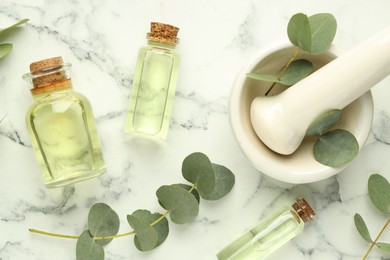 Photo of Bottles of essential oil, eucalyptus leaves, mortar and pestle on white marble table, flat lay