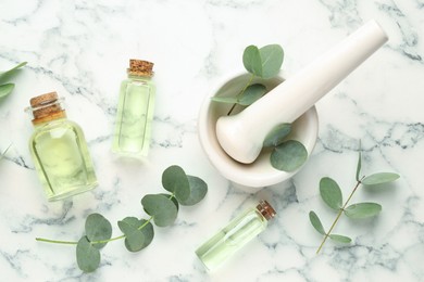 Photo of Bottles of essential oil, eucalyptus leaves, mortar and pestle on white marble table, flat lay