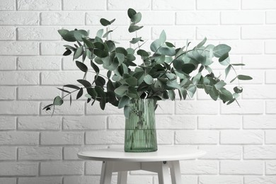 Photo of Green eucalyptus branches in vase on table near white brick wall indoors