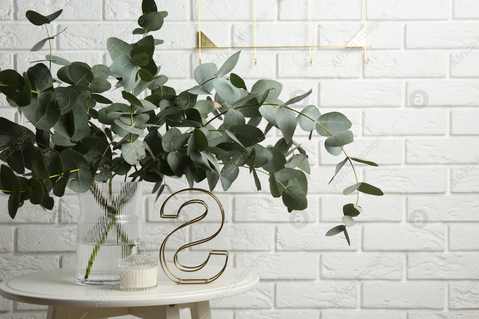Green eucalyptus branches in vase and other decor on table near white brick wall indoors Photo of Green eucalyptus branches in vase and other decor on table near white brick wall indoors
