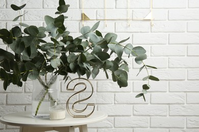 Photo of Green eucalyptus branches in vase and other decor on table near white brick wall indoors