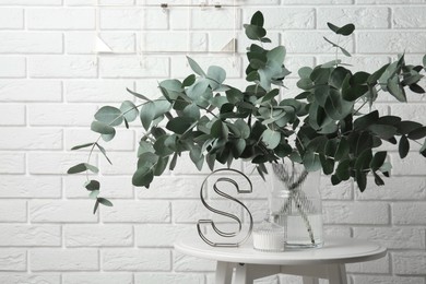 Photo of Green eucalyptus branches in vase and other decor on table near white brick wall indoors