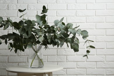 Photo of Green eucalyptus branches in vase on table near white brick wall indoors