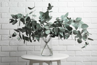 Photo of Green eucalyptus branches in vase on table near white brick wall indoors