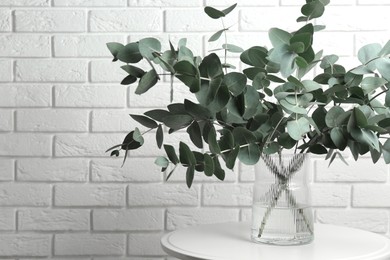 Photo of Green eucalyptus branches in vase on table near white brick wall indoors, space for text