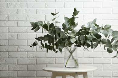 Photo of Green eucalyptus branches in vase on table near white brick wall indoors, space for text