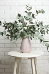 Photo of Green eucalyptus branches in vase on table near white brick wall indoors