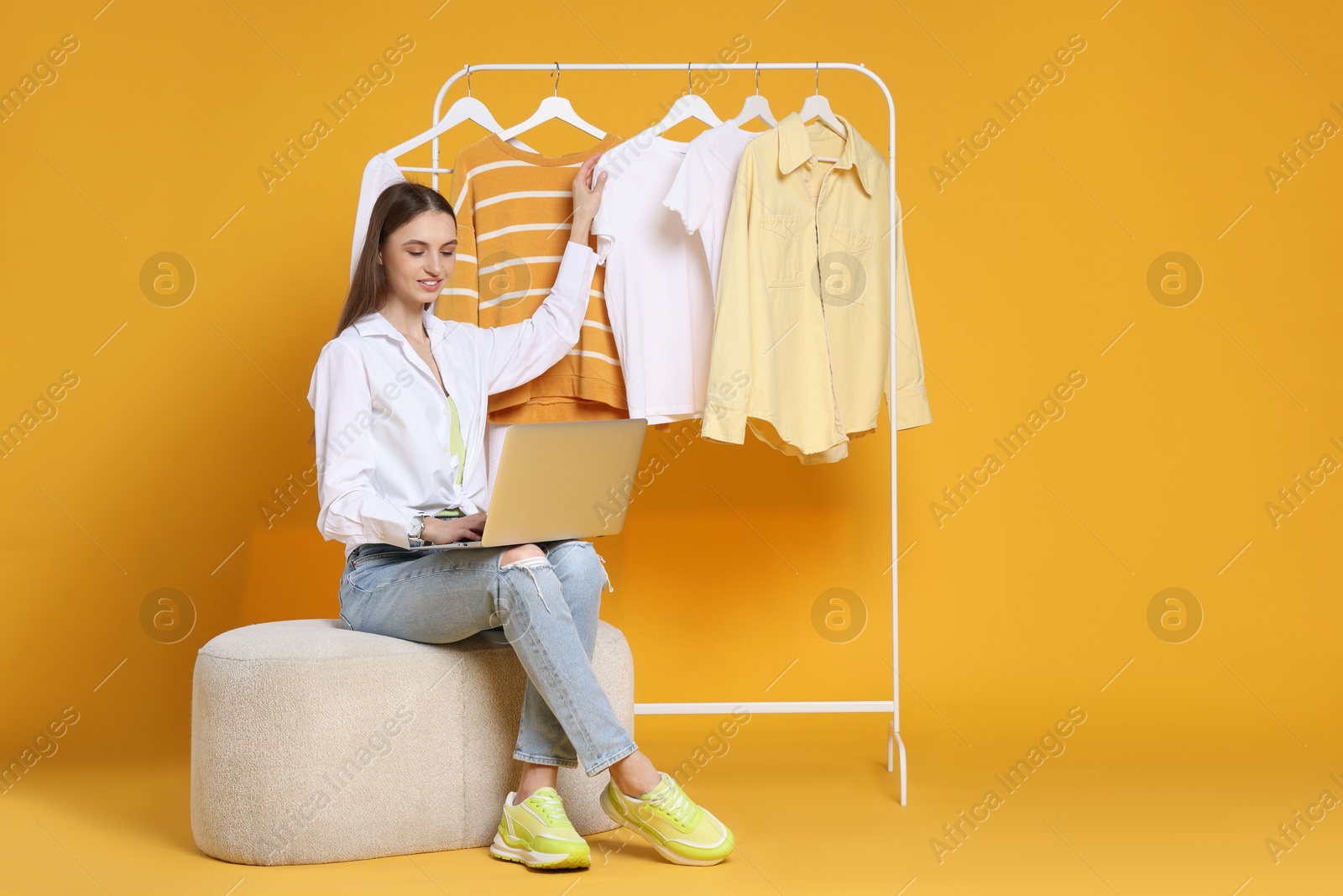 Smiling fashion stylist with laptop and clothing rack on yellow background Photo of Smiling fashion stylist with laptop and clothing rack on yellow background