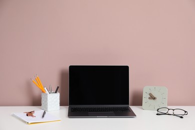 Photo of Laptop, different stationery, glasses and alarm clock on white office table against dark beige background
