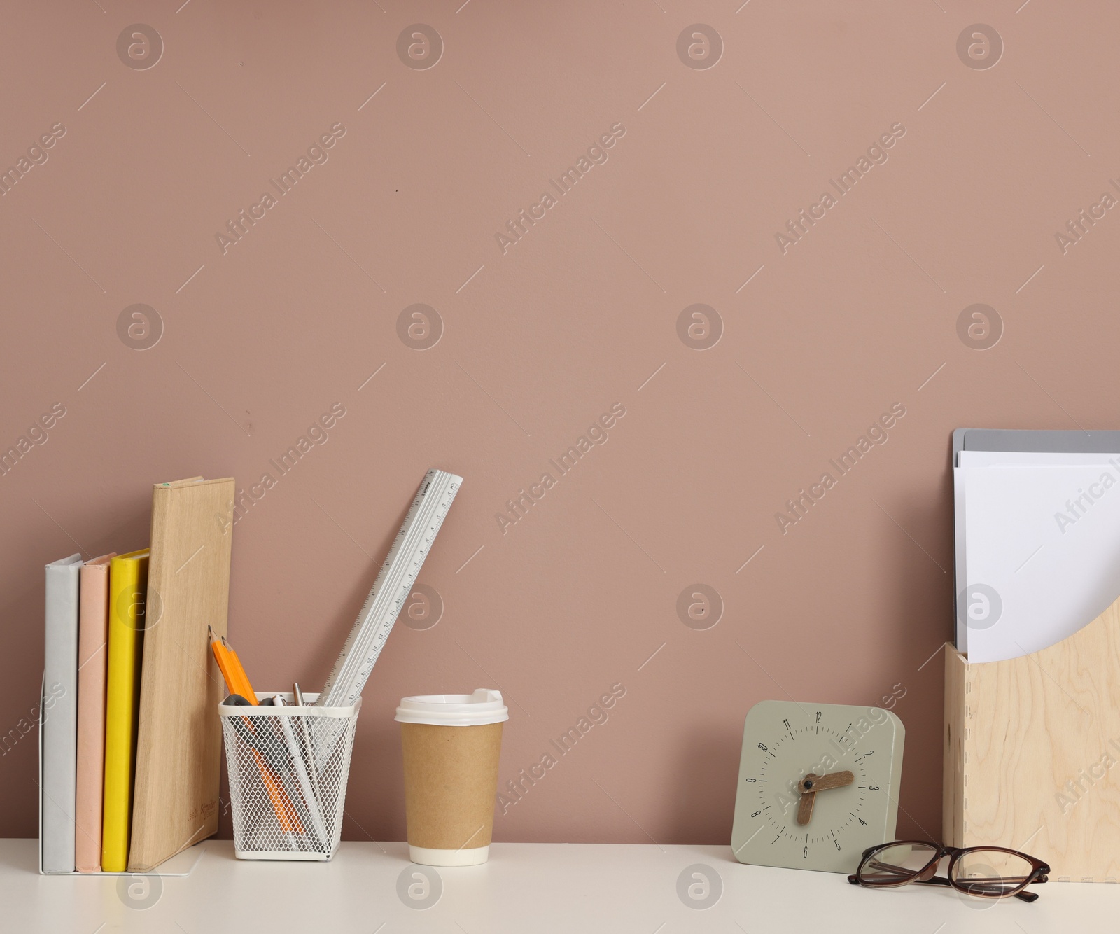 Different stationery, glasses and alarm clock on white office table against dark beige background Photo of Different stationery, glasses and alarm clock on white office table against dark beige background