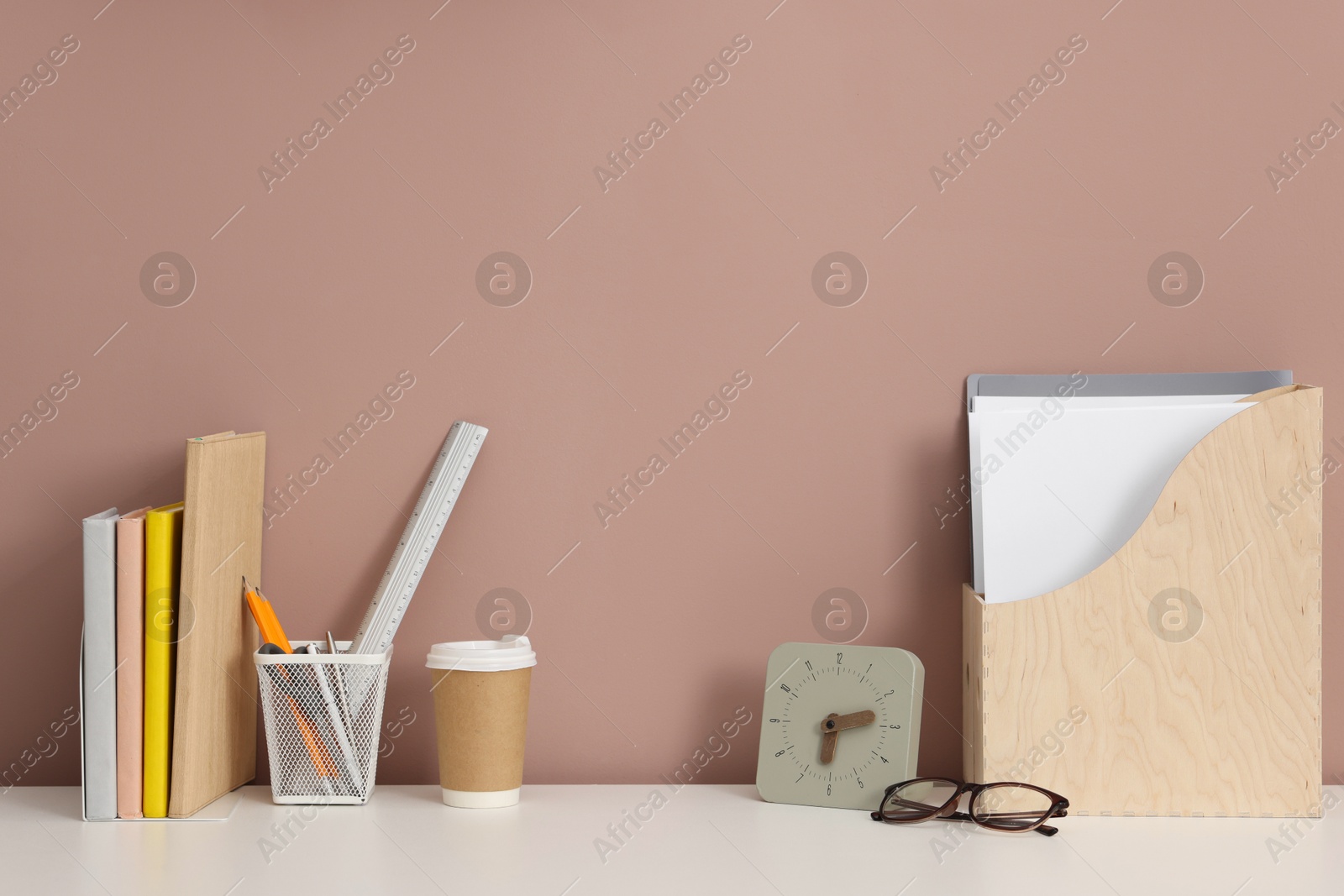 Different stationery, glasses and alarm clock on white office table against dark beige background Photo of Different stationery, glasses and alarm clock on white office table against dark beige background