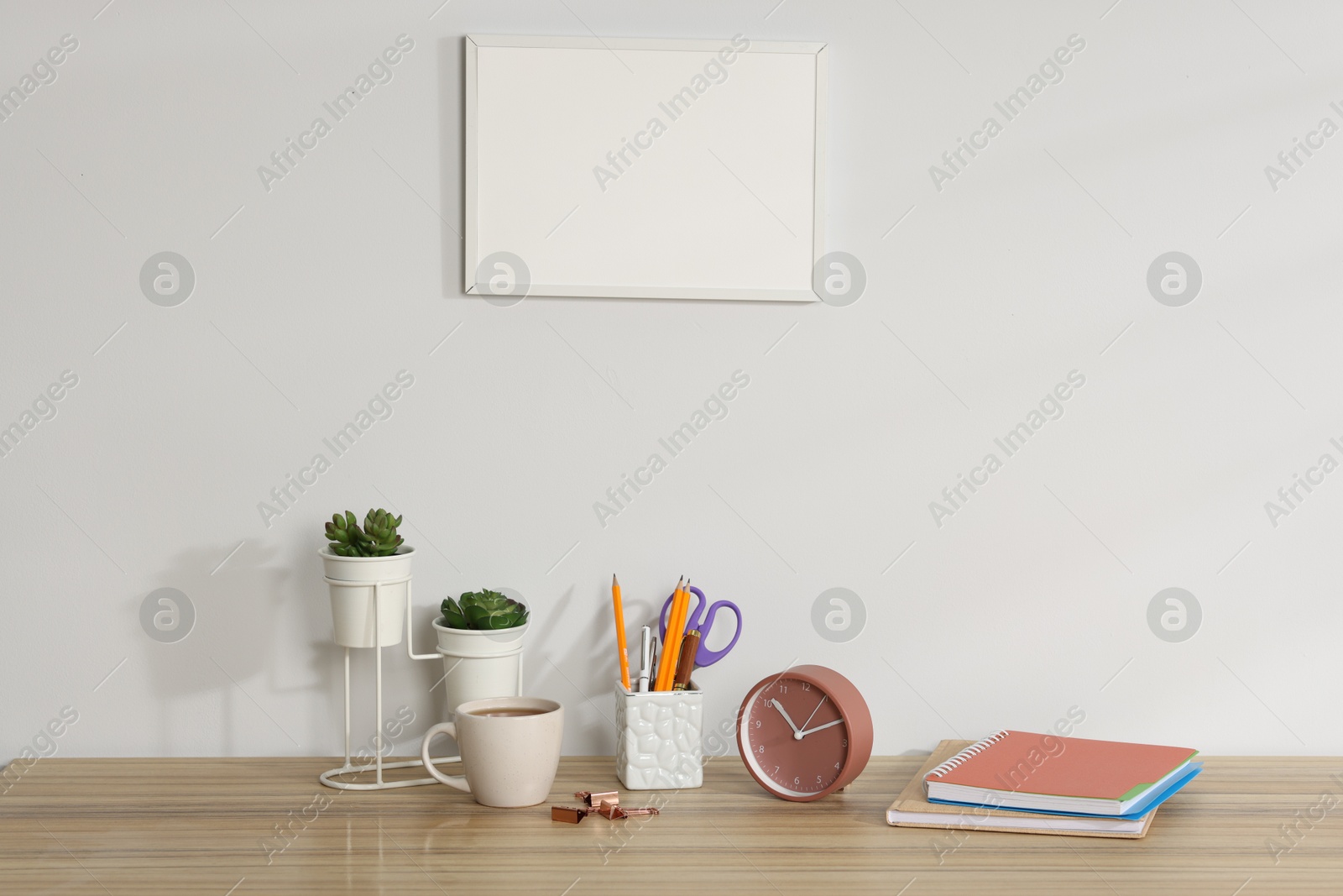 Different stationery, floral decor, alarm clock and coffee on wooden office table near white wall Photo of Different stationery, floral decor, alarm clock and coffee on wooden office table near white wall
