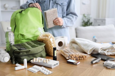 Photo of Survival kit. Woman packing different emergency supplies into backpack at wooden table indoors, selective focus