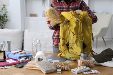 Photo of Survival kit. Woman putting raw pasta into backpack at wooden table indoors, closeup