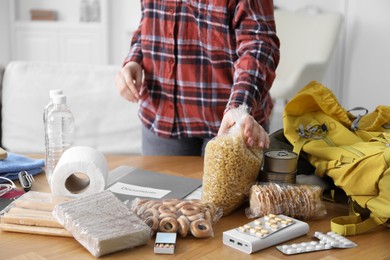 Photo of Survival kit. Woman packing different emergency supplies at wooden table indoors, closeup