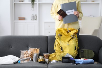 Photo of Survival kit. Woman holding passport and documents near sofa with different emergency supplies indoors, closeup
