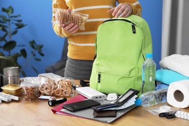 Photo of Survival kit. Woman packing different emergency supplies into backpack at wooden table indoors, closeup