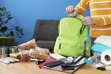 Photo of Survival kit. Woman packing different emergency supplies into backpack at wooden table indoors, closeup