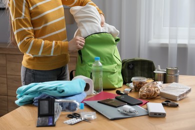 Photo of Survival kit. Woman packing different emergency supplies into backpack at wooden table indoors, closeup