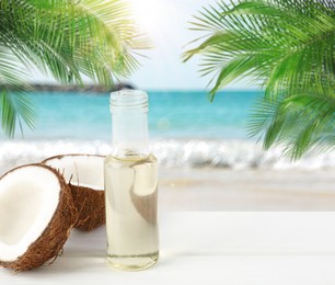 Image of Cooking oil in glass bottle and coconut on white table against beach. Space for text