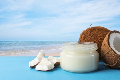 Image of Cooking oil in jar and coconuts on blue table against beach. Space for text