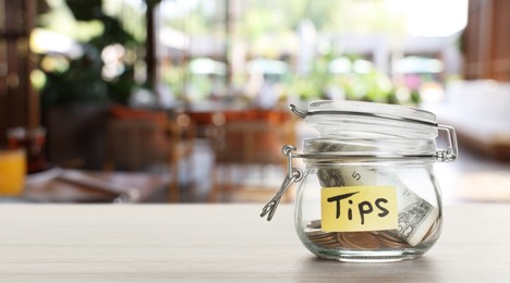 Image of Tips jar with money on light wooden table in cafe, space for text. Banner