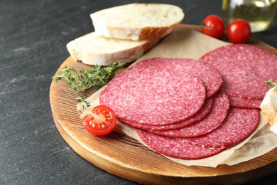 Photo of Slices of tasty smoked sausage, tomatoes, thyme and bread on black table, closeup