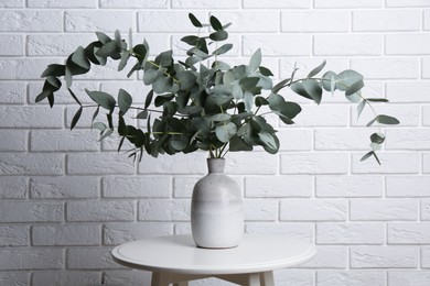 Photo of Green eucalyptus branches in vase on table near white brick wall indoors