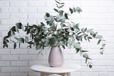 Photo of Green eucalyptus branches in vase on table near white brick wall indoors