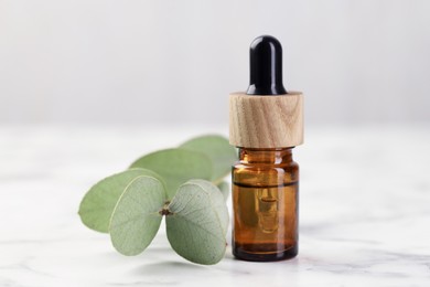 Photo of Bottle of essential oil and eucalyptus leaves on white marble table, closeup