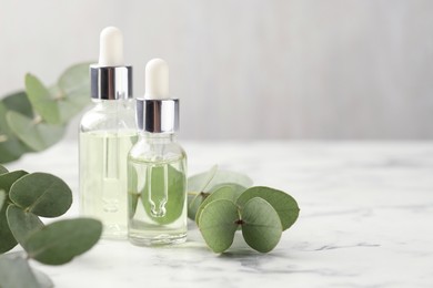 Photo of Bottles of essential oil and eucalyptus leaves on white marble table, closeup. Space for text