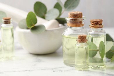Photo of Bottles of essential oil, eucalyptus leaves, mortar and pestle on white marble table, closeup