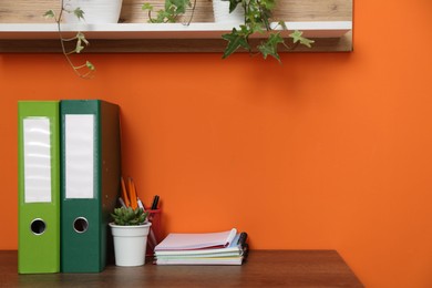 Photo of Stylish workplace with stationery, folders and plant on wooden desk near orange wall. Space for text