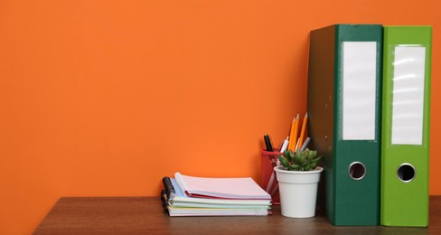 Photo of Stylish workplace with stationery, folders and plant on wooden desk near orange wall. Space for text