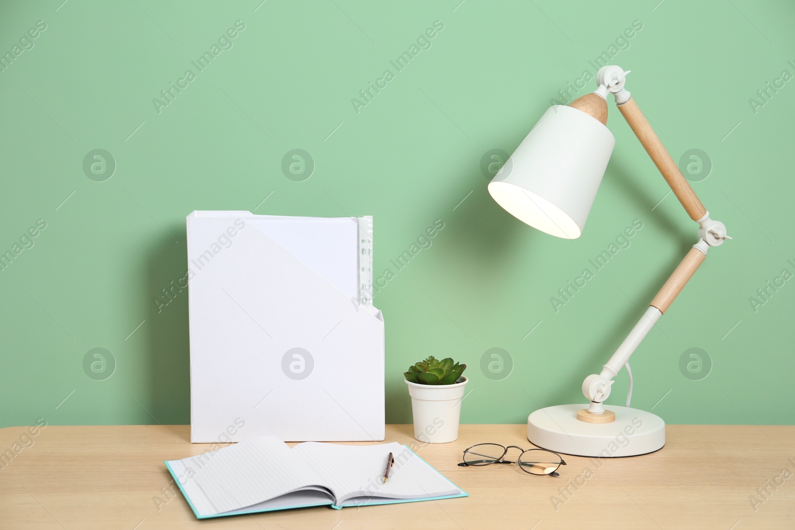 Stylish workplace with lamp, stationery and plant on wooden desk near light green wall Photo of Stylish workplace with lamp, stationery and plant on wooden desk near light green wall