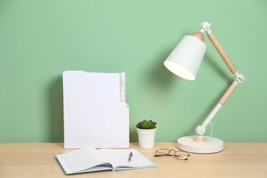 Photo of Stylish workplace with lamp, stationery and plant on wooden desk near light green wall