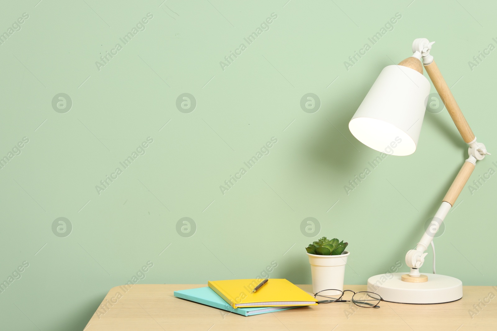 Stylish workplace with lamp, stationery and plant on wooden desk near light green wall. Space for text Photo of Stylish workplace with lamp, stationery and plant on wooden desk near light green wall. Space for text