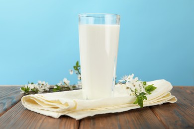 Photo of Fresh milk in glass and blossoms on wooden table against light blue background, closeup