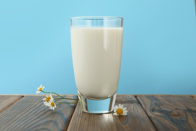 Photo of Fresh milk in glass and flowers on wooden table against light blue background, closeup