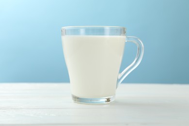 Photo of Fresh milk in glass cup on white wooden table against light blue background, closeup