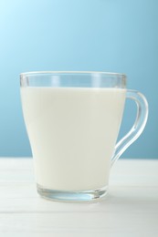 Photo of Fresh milk in glass cup on white wooden table against light blue background, closeup